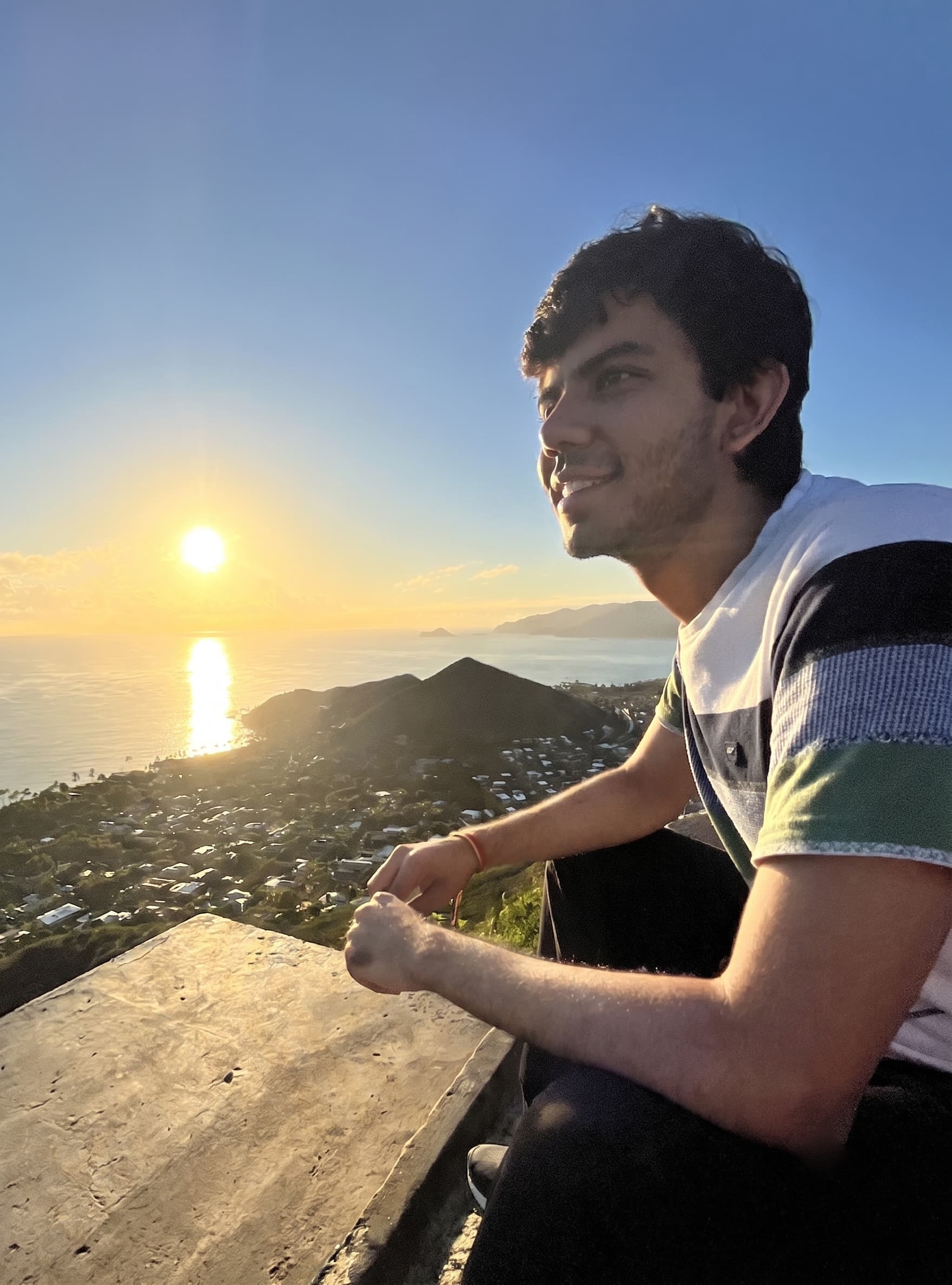 Utkarsh Bali sitting on a hilltop above the coast, smiling into the light as the sun drops over the ocean behind him